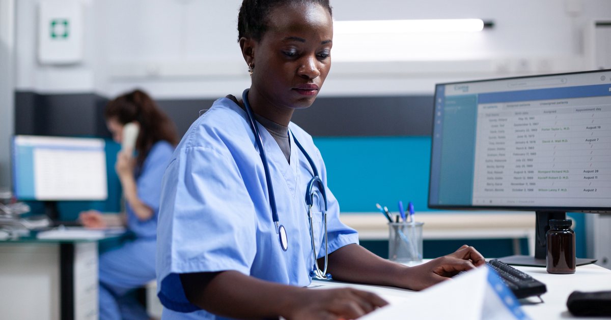 A nurse checking a paper document against a patient list on her monitor