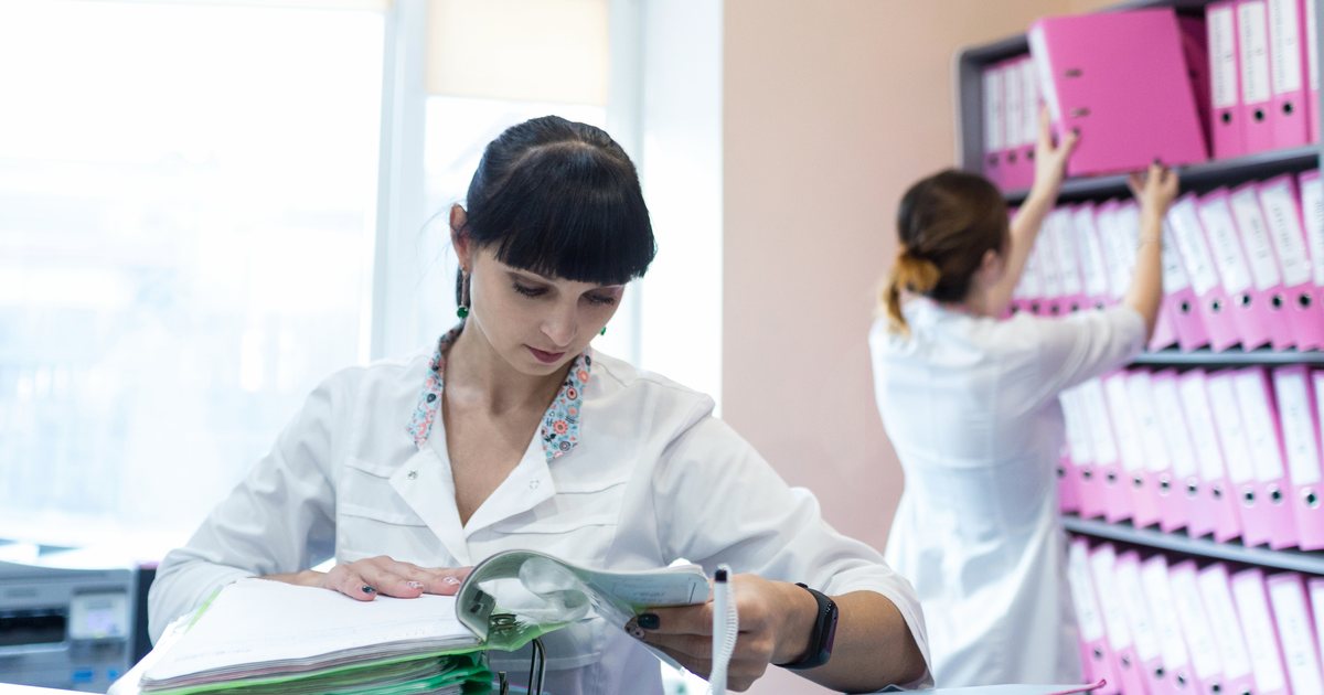A staff member working through a thick stack of patient folders while a colleague files binders behind her