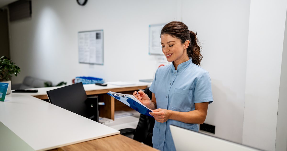 A medical assistant at the front desk reading a paper form on a clipboard